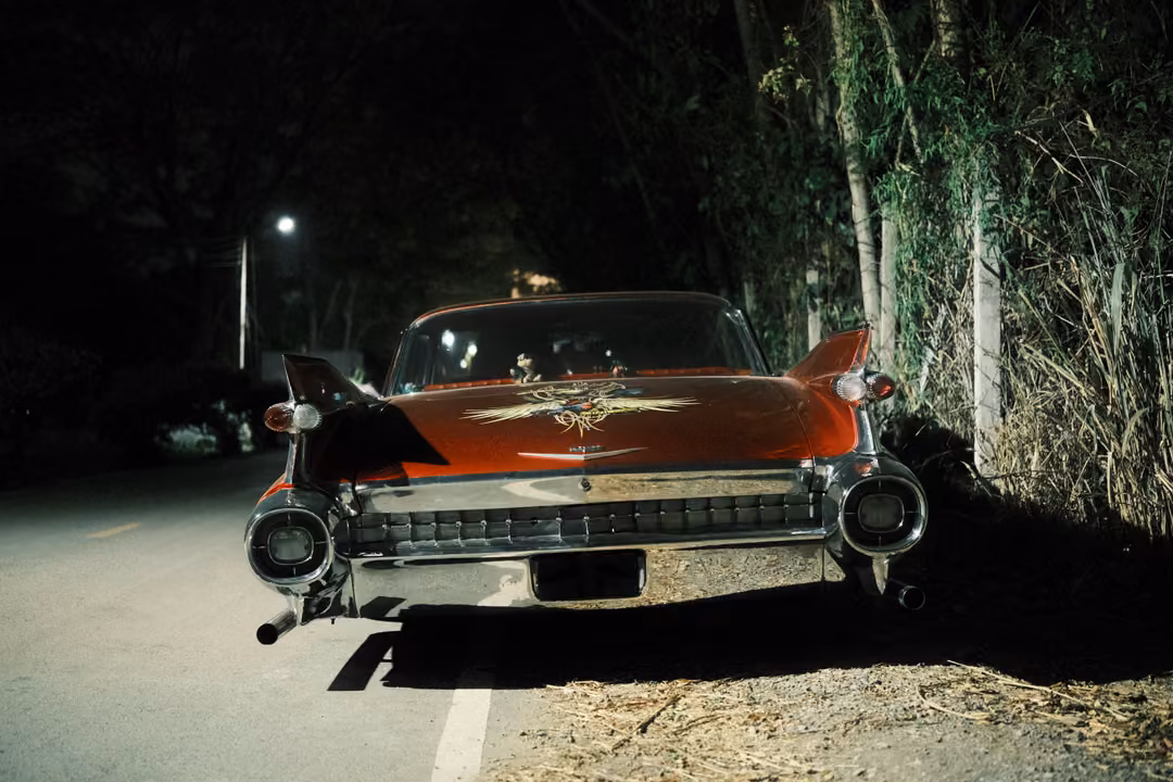 Rear of a red vintage car with tall tail fins and twin round lights parked by trees on a dark roadside