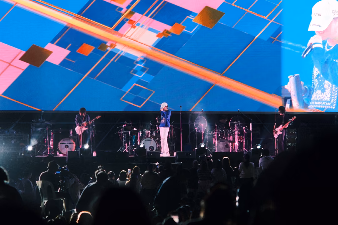 Band on stage below blue and pink geometric graphics with an orange diagonal streak, singer close-up at screen edge.