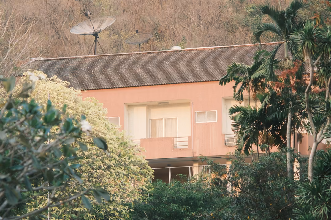 Pink two-storey building with a tiled roof and satellite dishes, surrounded by palms and trees, dry hill behind