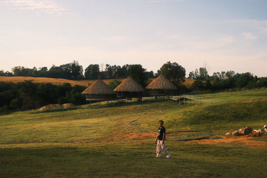 A person walks across a sunlit green lawn toward the foreground, thatched huts on a low hill behind.