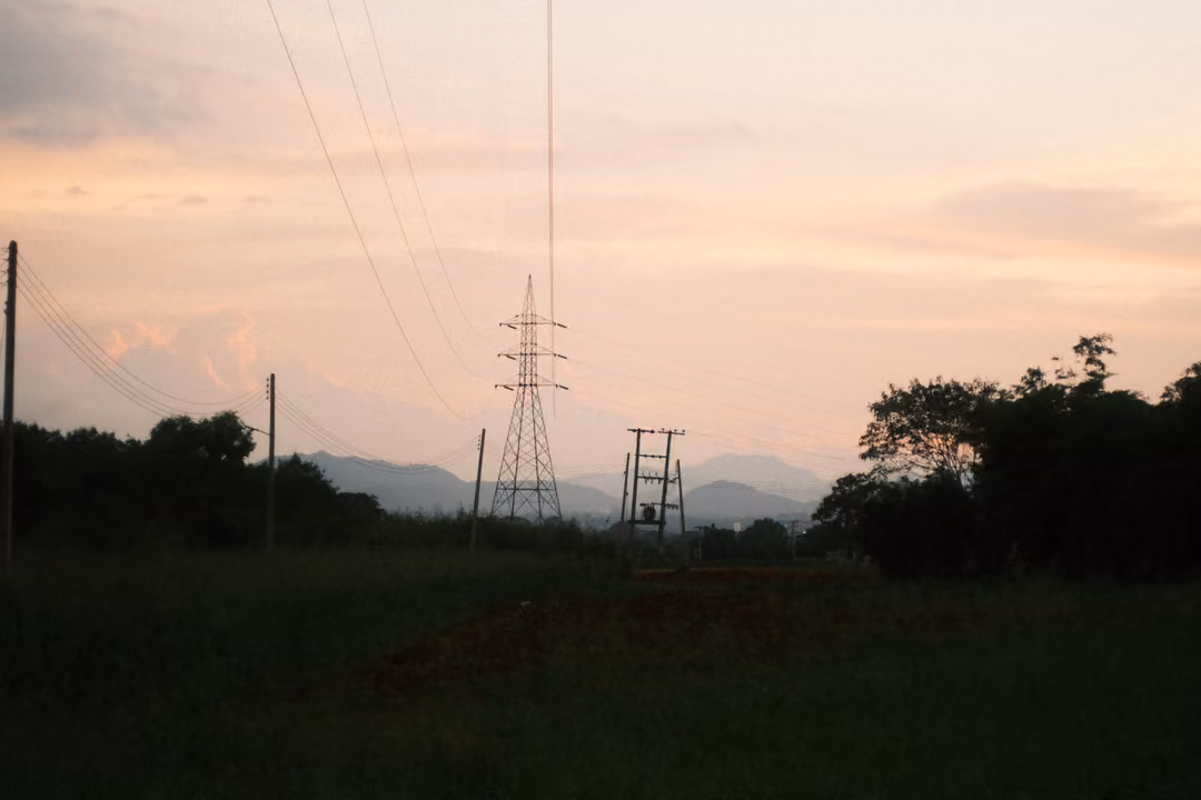 Electricity pylon and distant mountains against a pink cloudy evening sky, dark field below