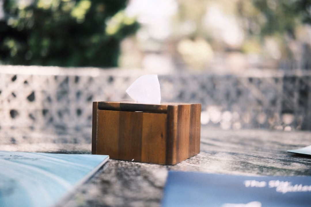 A wooden tissue box on a dark table beside menus, a patterned concrete wall and greenery blurred behind