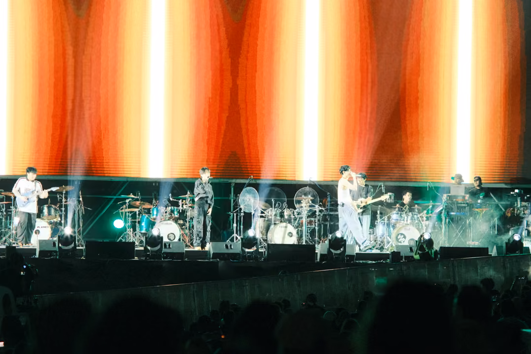 Band on a stage lit by an orange and red striped screen, singer and guitarist together at right, haze drifting.