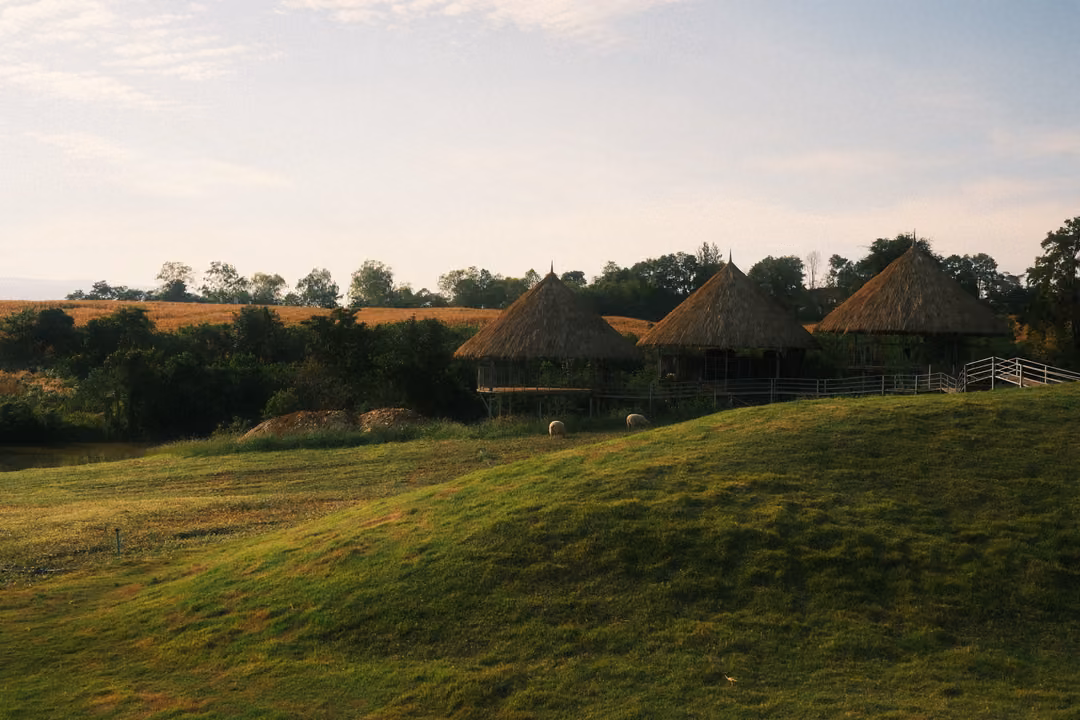 Thatched huts on stilts beside trees, beyond a rolling green mound under a pale blue sky.