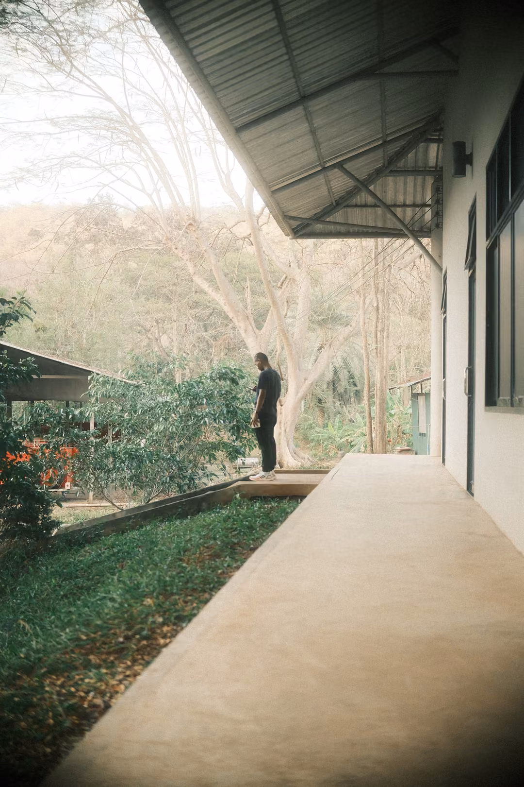 Covered concrete walkway alongside a white building, a man standing at the far end, hazy trees beyond