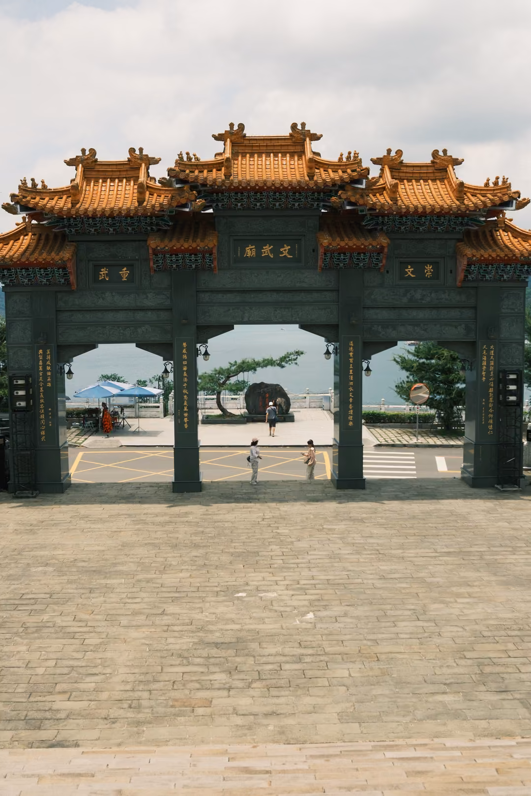 Tall ornamental gateway with golden tiled roofs over a stone plaza, lake and trees visible through the arches
