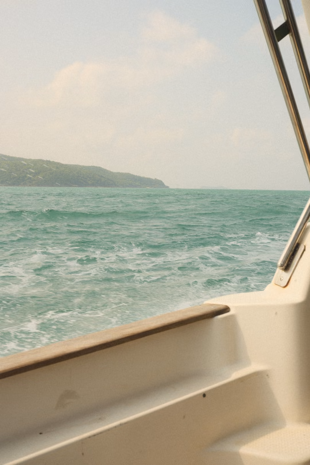 From the boat's stern corner, foamy turquoise water and a long green headland on the horizon.
