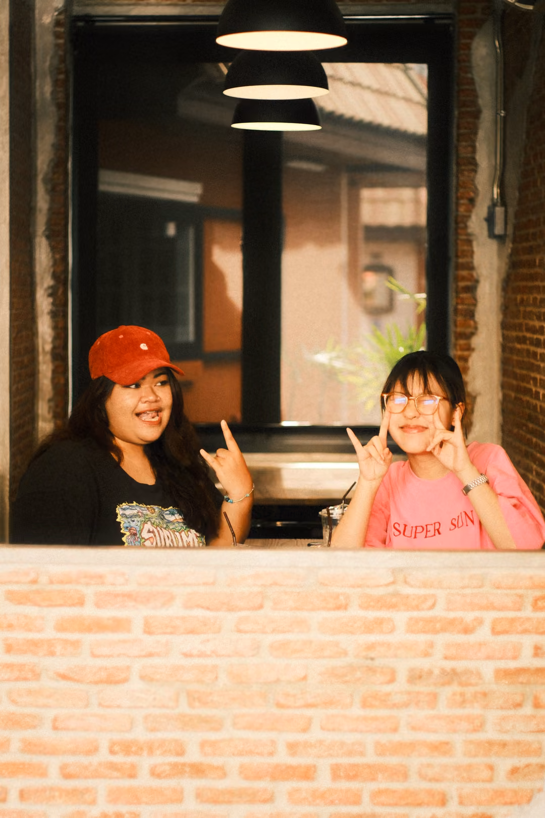 Two women posing at a window behind a brick wall, one in a red cap, the other in pink making hand gestures