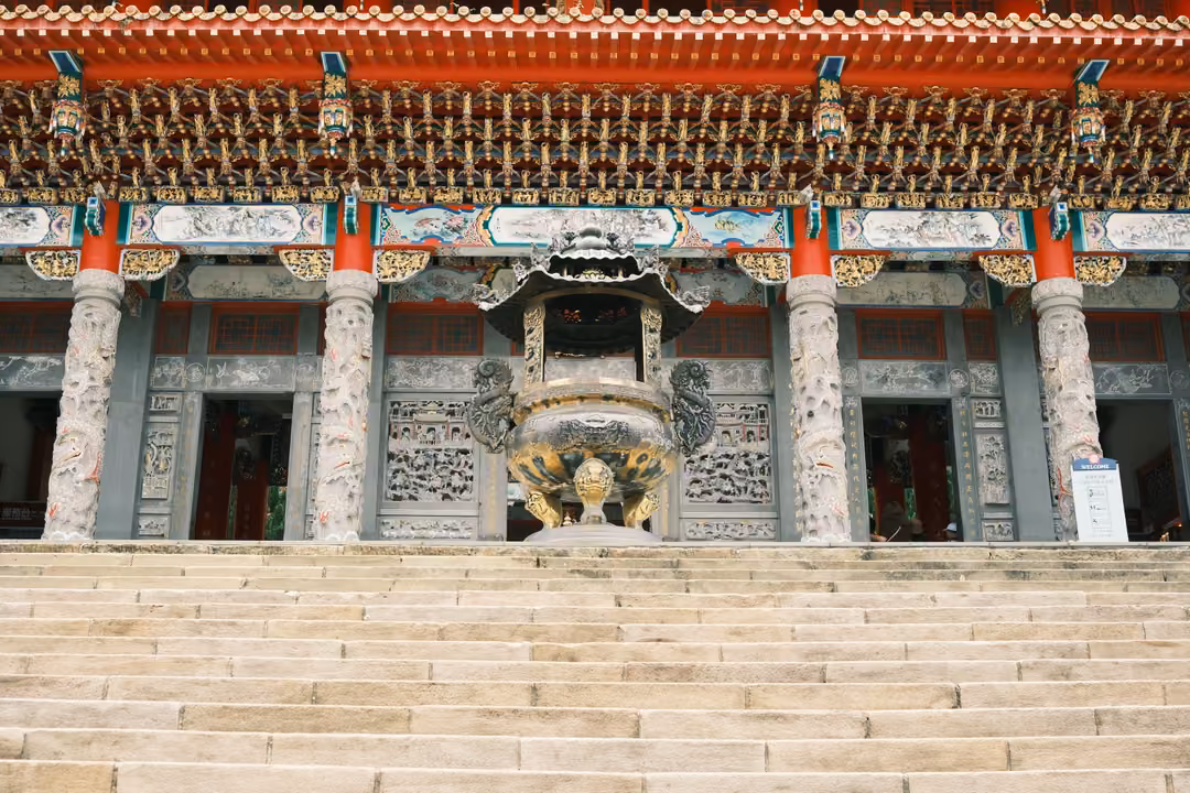 Front of an ornate temple with red roof eaves, carved stone columns and a bronze incense burner atop wide steps