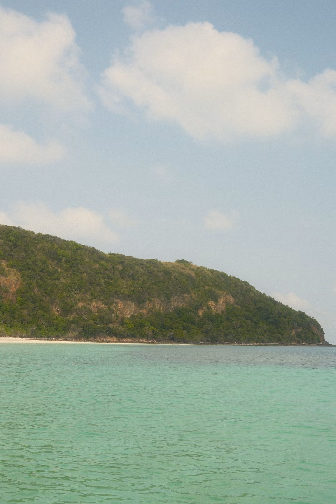 A green headland sloping into turquoise sea under a blue sky with puffy white clouds.
