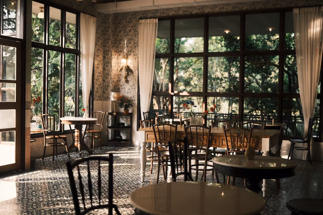 Bright café room with tall black-framed windows, white curtains, patterned tile floor and round tables