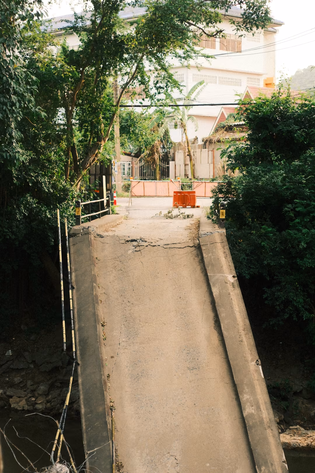 A broken concrete bridge deck tilting down over a stream, leading up to an orange barrier and buildings beyond.