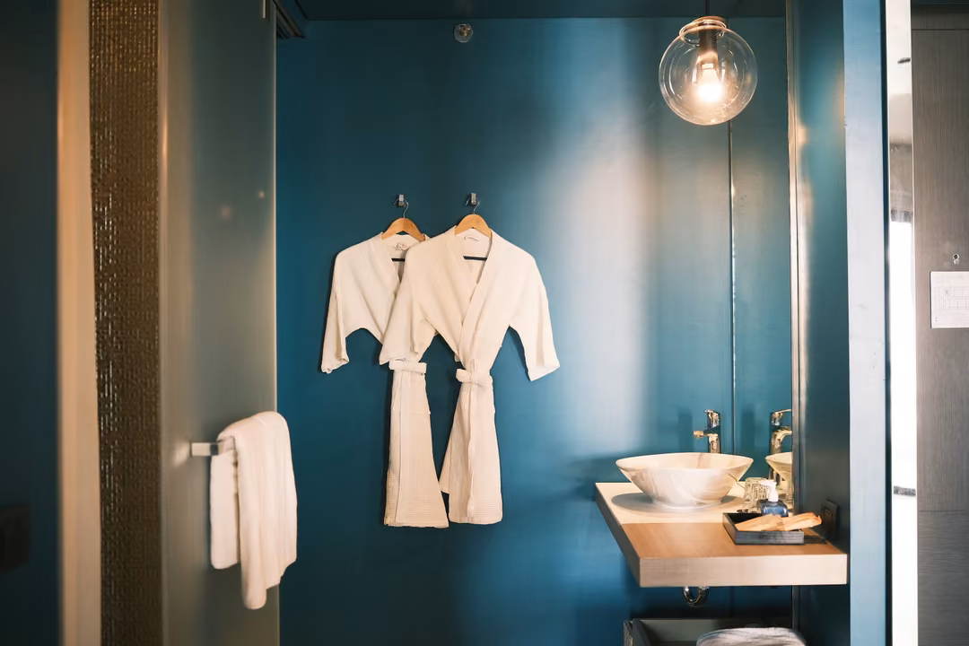 A teal bathroom with two white robes, a hanging bulb, a basin on a wooden shelf and a towel rail