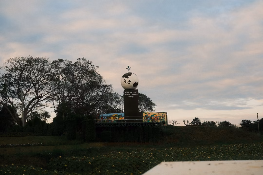 White globe sculpture on a dark pillar beside large trees, a colourful mural wall and an open field under clouds