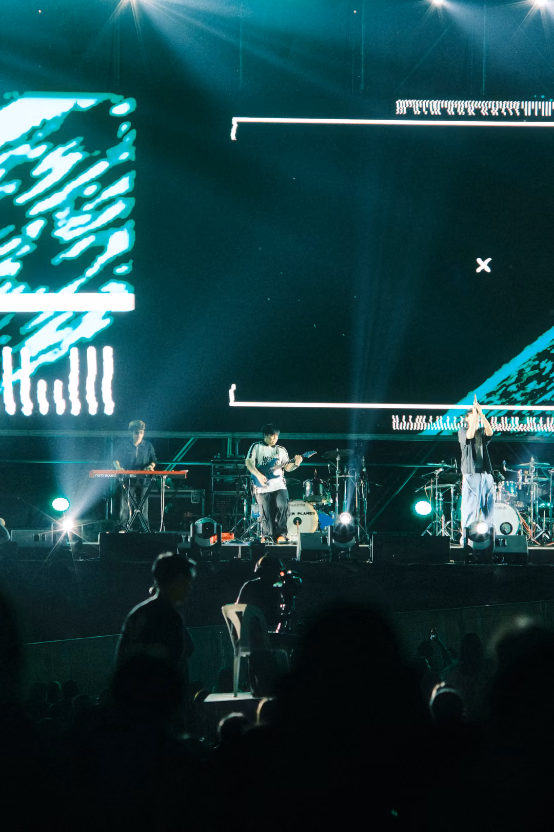 Vertical view of the stage with a white bracket and cross on the screen, green streaks at left, band and crowd below.