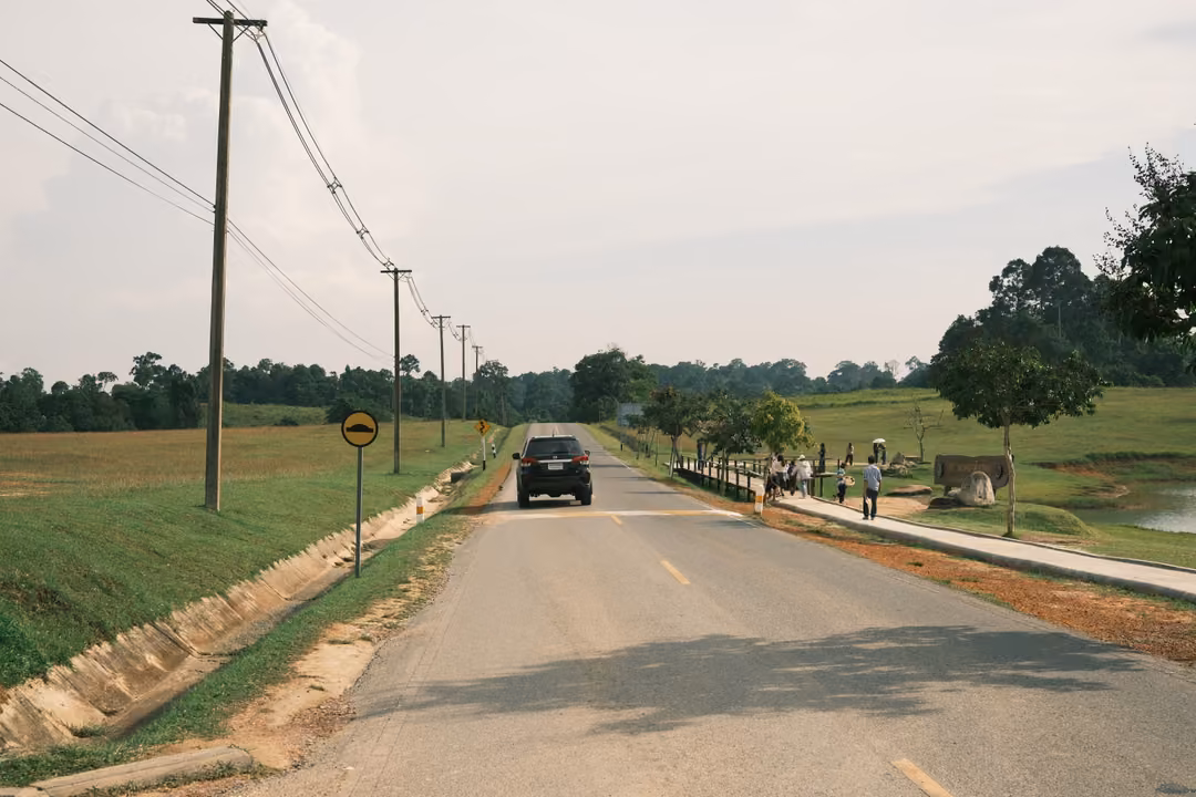 A straight road between grassy verges and power poles, with a dark SUV approaching and walkers on a footpath.