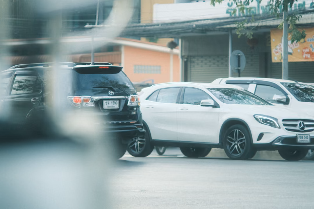 A black SUV and a white Mercedes parked in front of shophouses, seen past a blurred pale shape in the foreground.