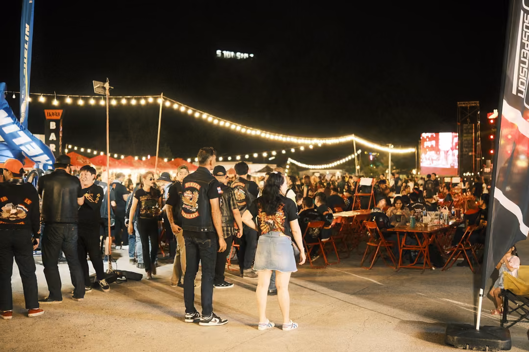 Groups of people in dark clothes standing near flags at a night festival, tables and string lights behind