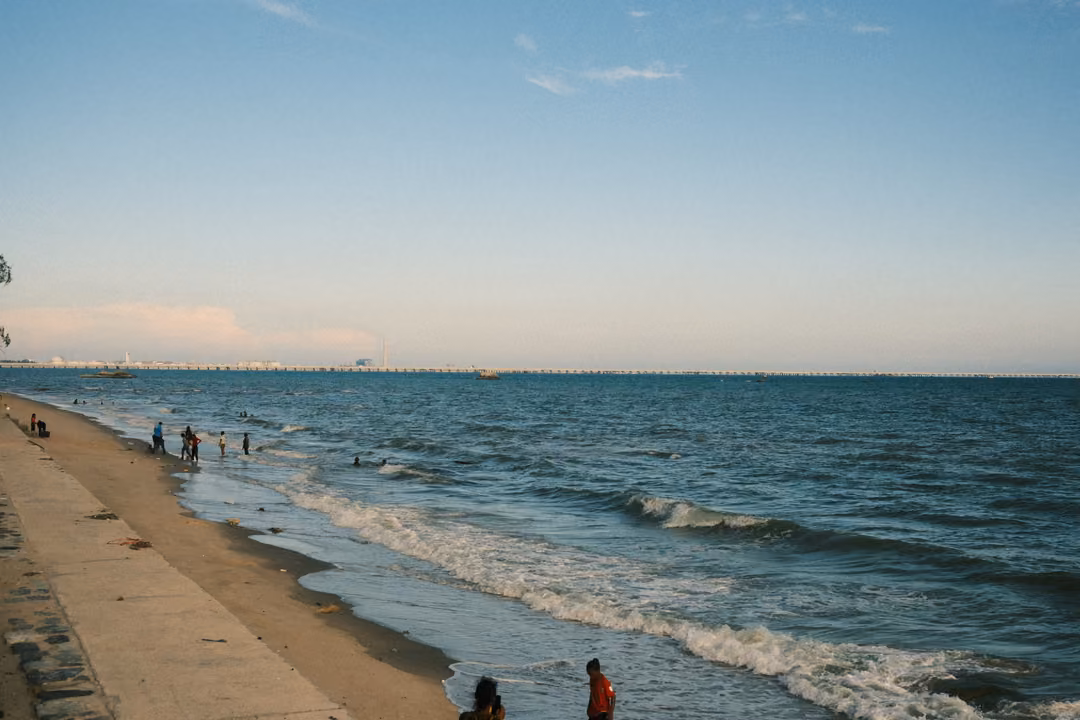 Seawall and sandy beach curving into the distance with people in the waves and a long pier on the horizon