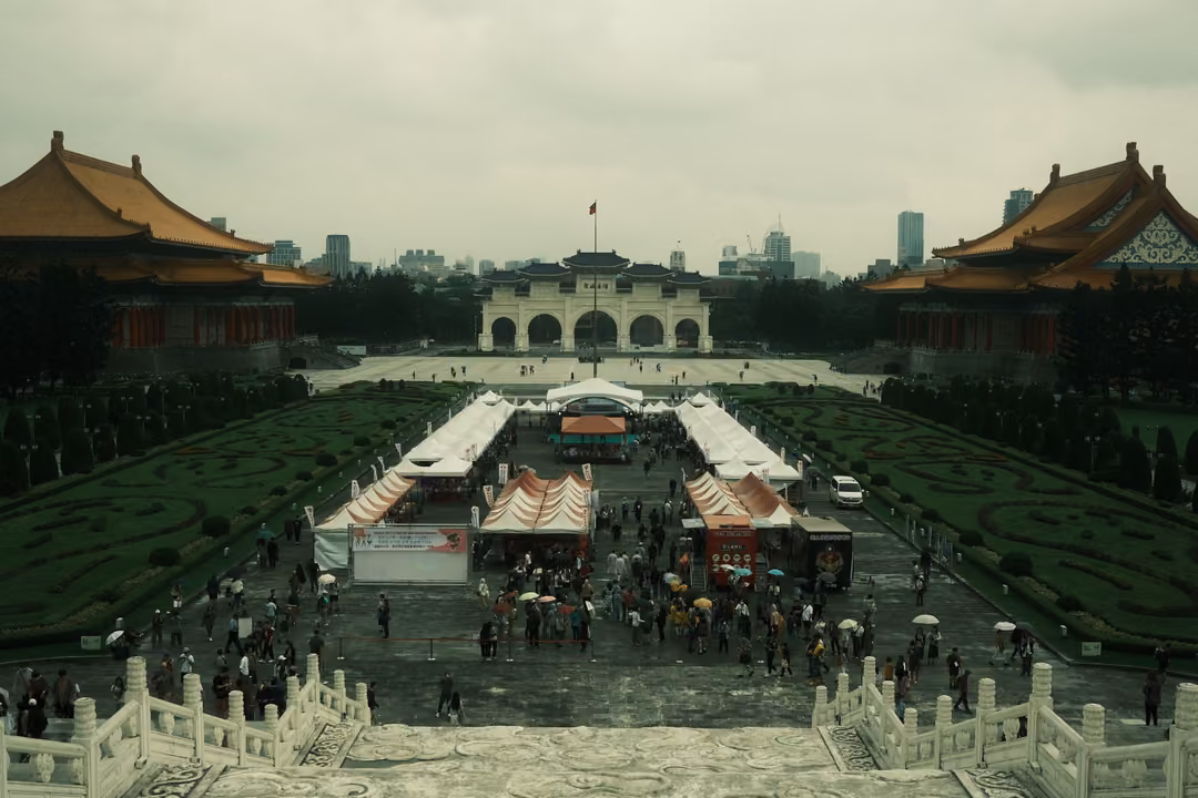 Same plaza view, white tents and umbrella-carrying crowd between two traditional halls under overcast sky