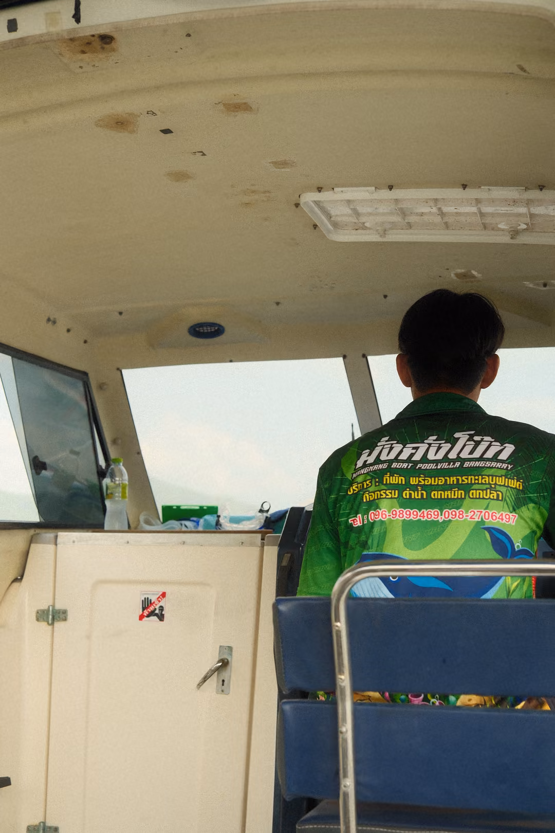 Inside a boat cabin, a driver in a bright green printed shirt sits at the wheel, facing forward.