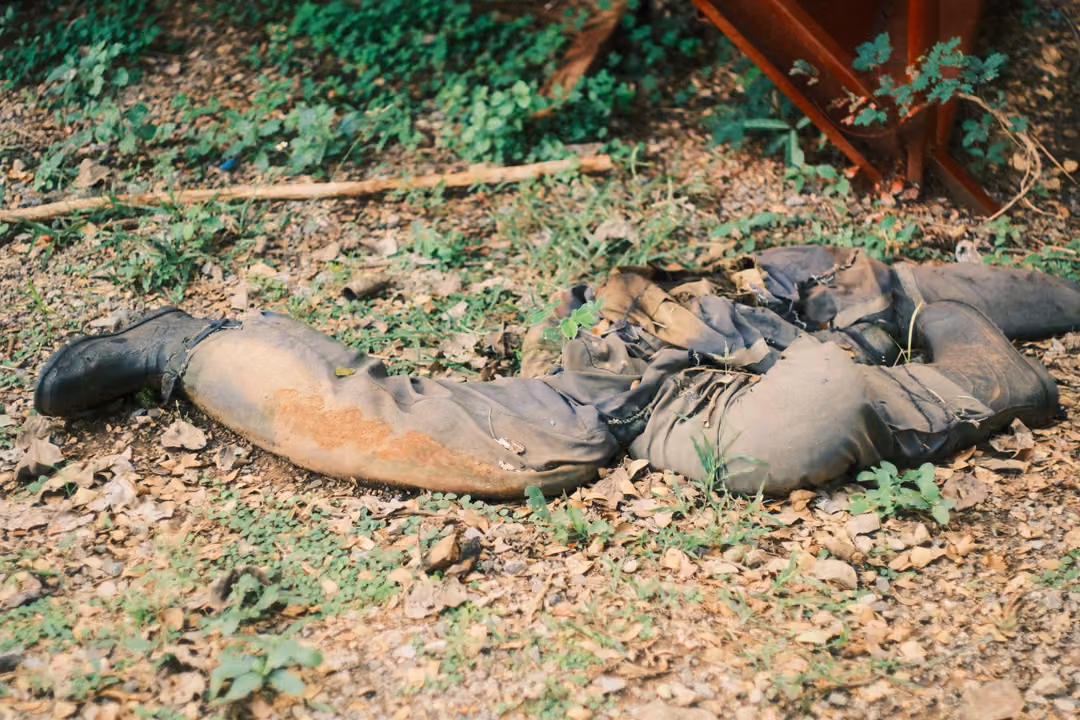 Muddy fatigues and black boots lying empty on dry leaves and weeds beside a rusty red structure