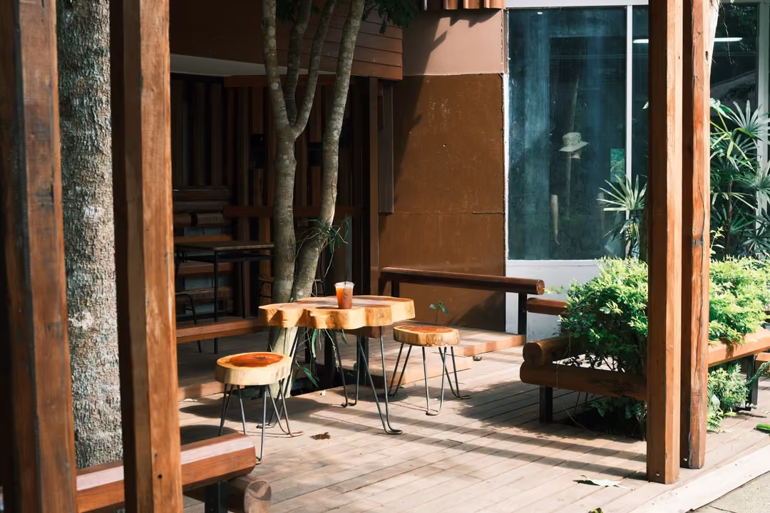 A wooden café terrace with tree-trunk tables and stools, a drink on the table, plants and glass behind