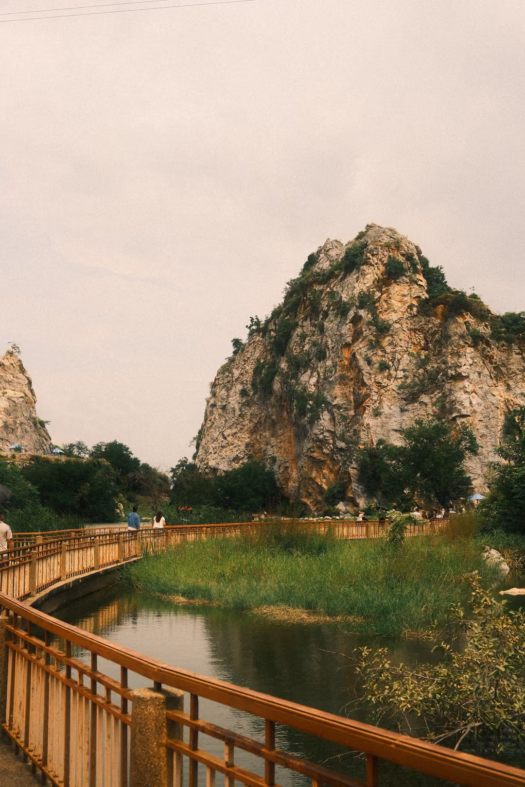 Wooden railed boardwalk curving across a marshy lake toward a tall limestone peak under a pale sky