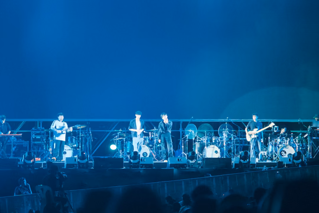 Band performing on a stage under a plain blue screen, a soft green glow at the right, crowd silhouetted.