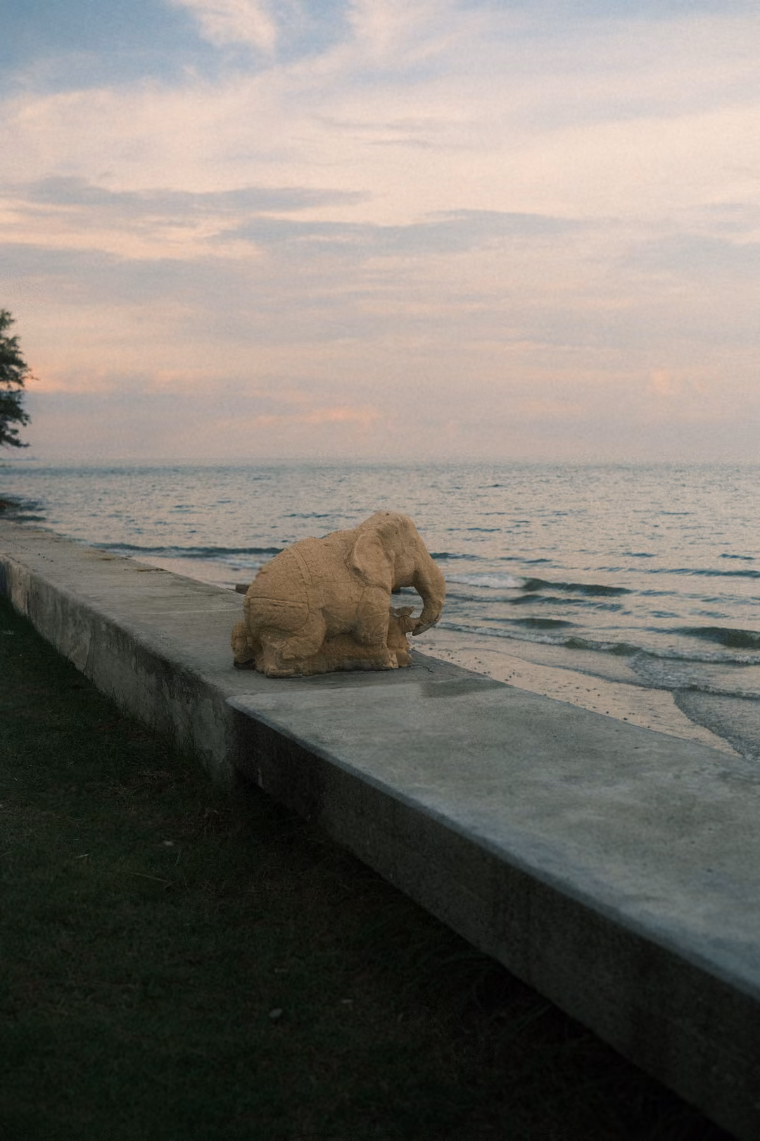 Vertical frame of a carved stone elephant sitting on a seawall above the beach, sea and pink sky behind