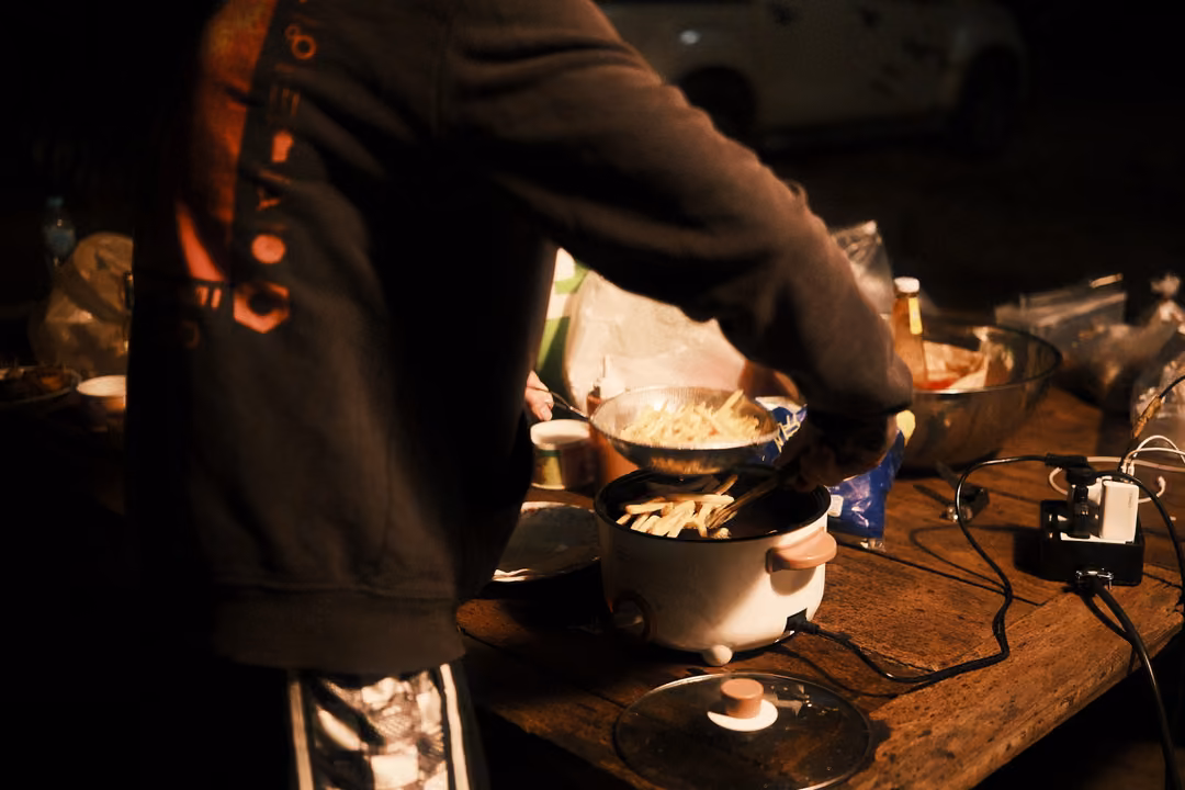 Person in a dark jacket stirring food in a pot on a rice cooker at a camp table at night