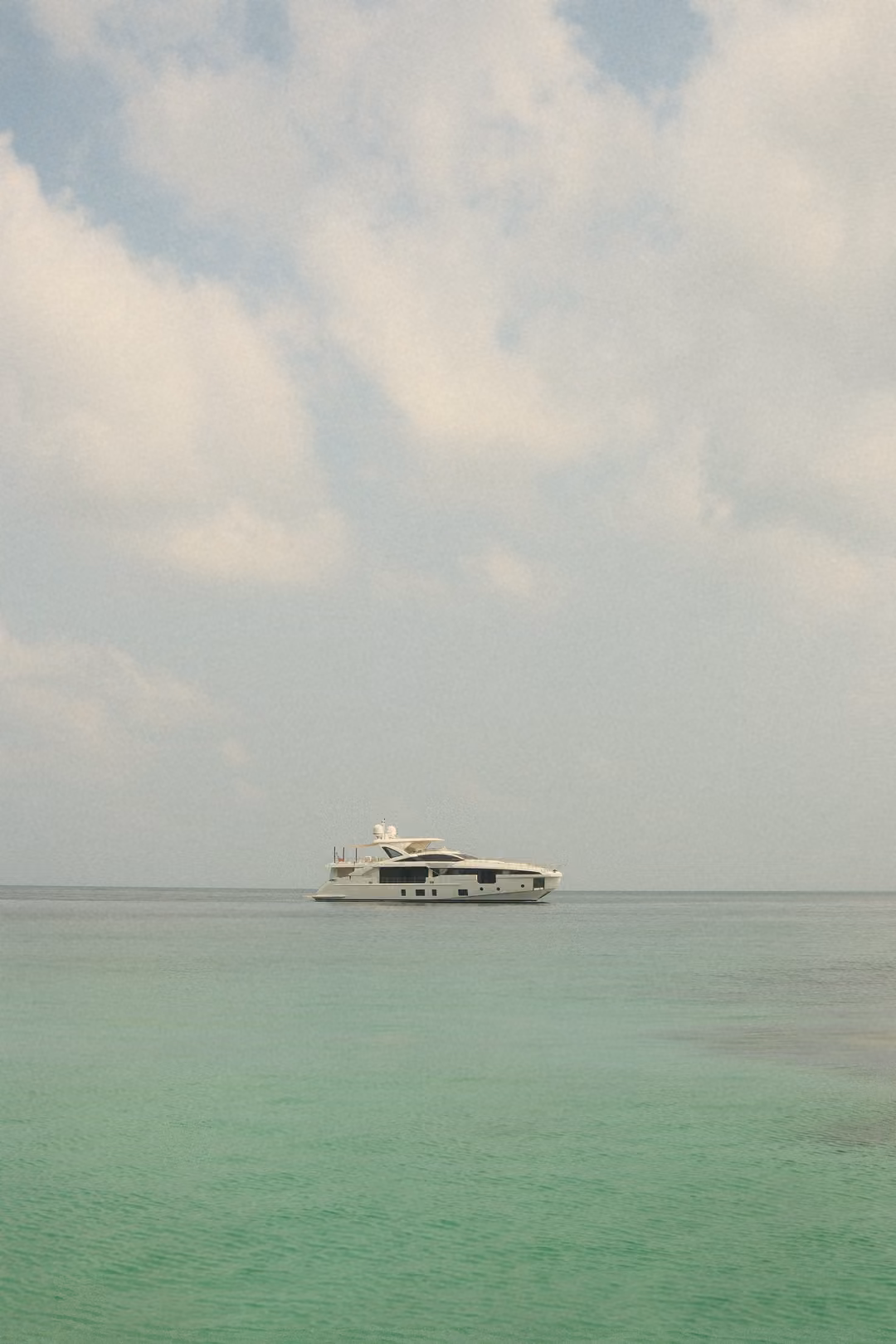 A white yacht anchored far off on pale green sea beneath a soft, cloud-filled sky.