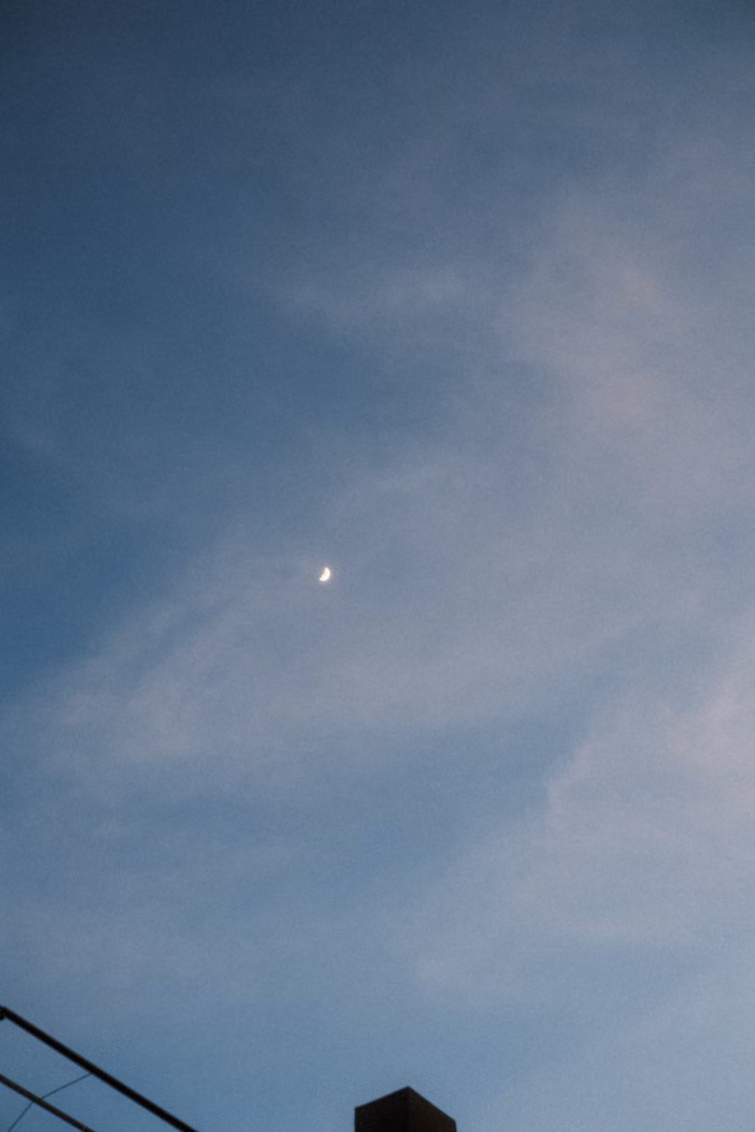 Thin crescent moon in a dusky blue sky with wispy clouds, a wire and post at the bottom
