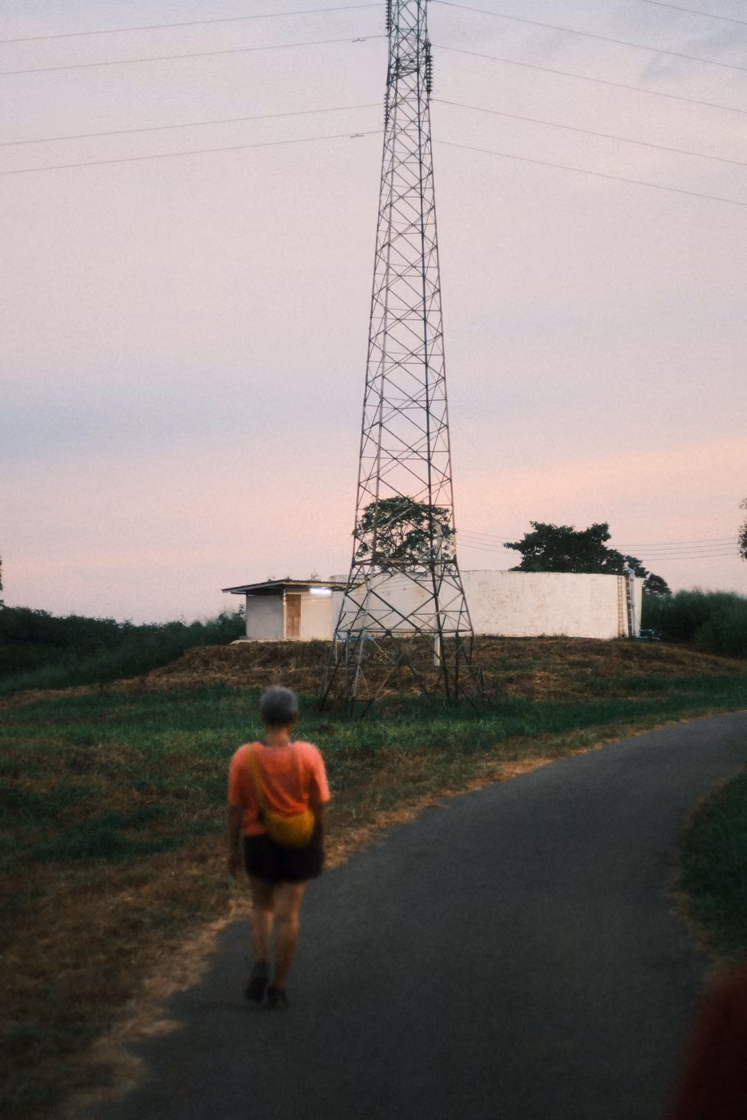 Person walking away down a narrow path toward a tall electricity pylon and a small white building