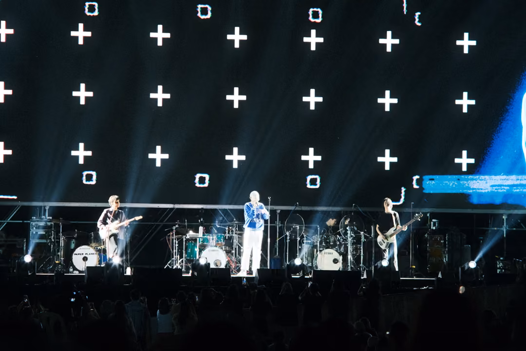Band on stage below a screen showing a grid of white plus signs and squares, beams of light crossing the haze.
