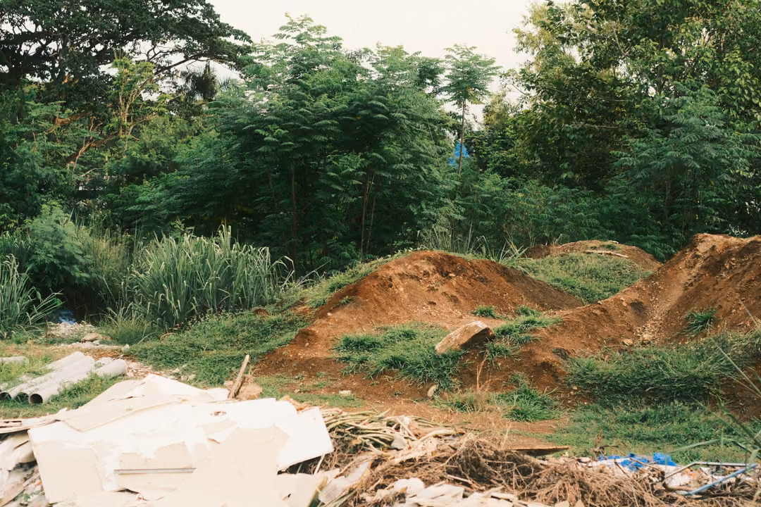 Mounds of freshly dug red earth beside broken white rubble and debris, with green trees behind.