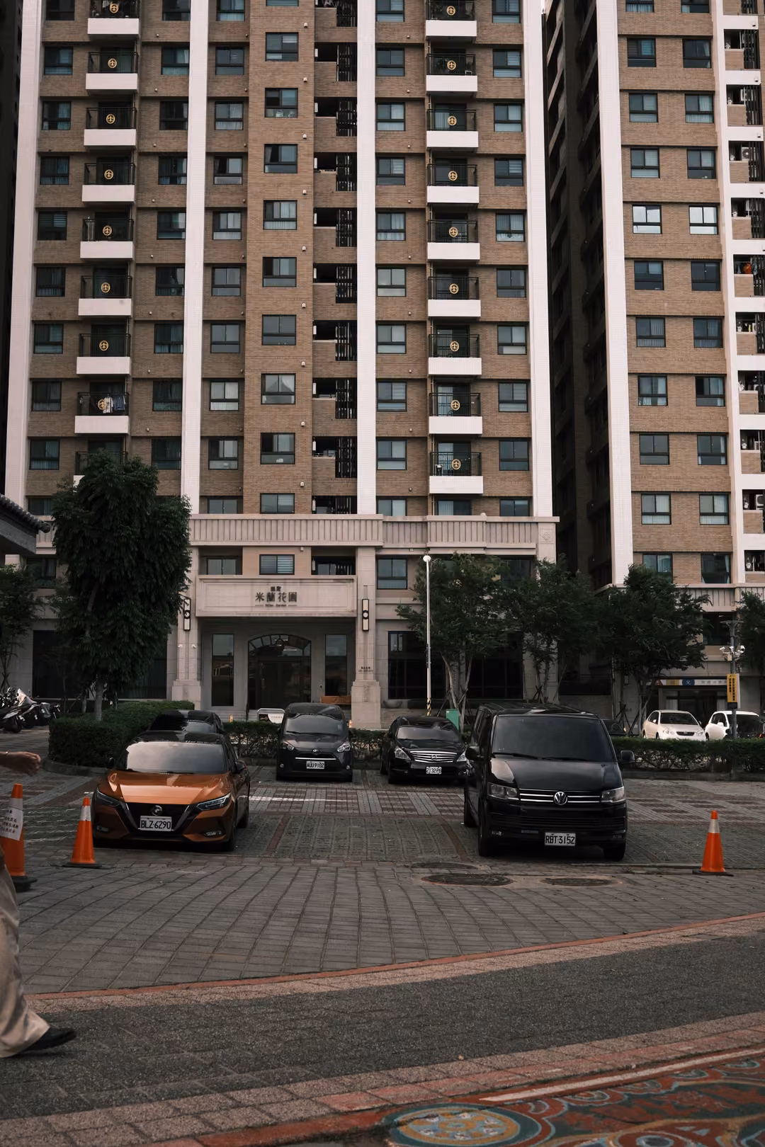Vertical view of residential towers above a parking area with orange cones, a passing leg at the left edge