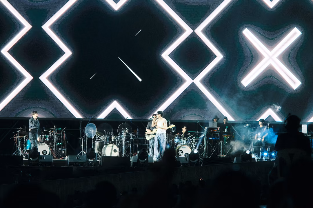 Band on stage under a black screen patterned with large white X shapes and a thin streak of light.