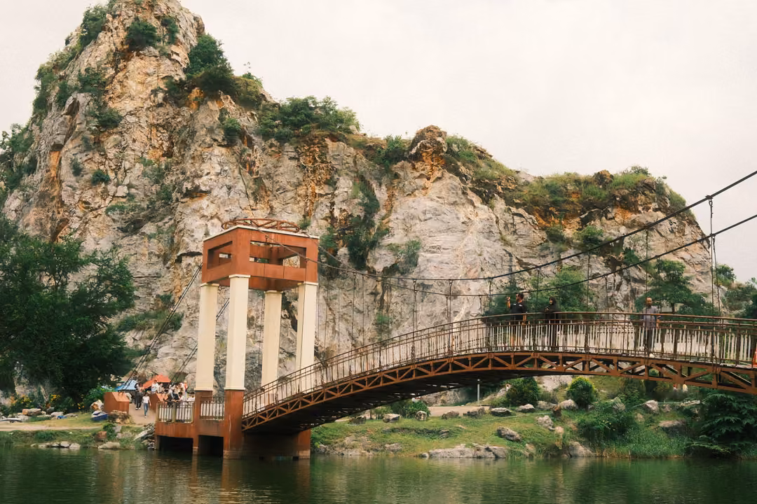 Arched suspension bridge with a red and white tower crossing a lake toward a limestone cliff, people on the bridge