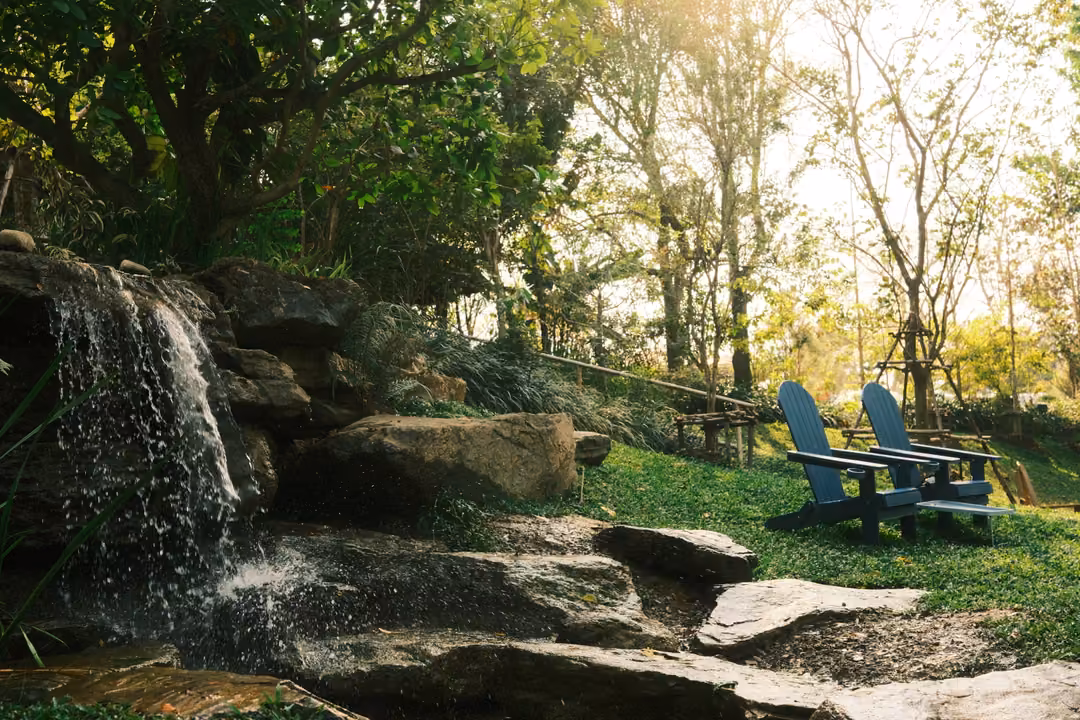 Small waterfall tumbling over rocks beside a lawn with two blue wooden lounge chairs in afternoon sun