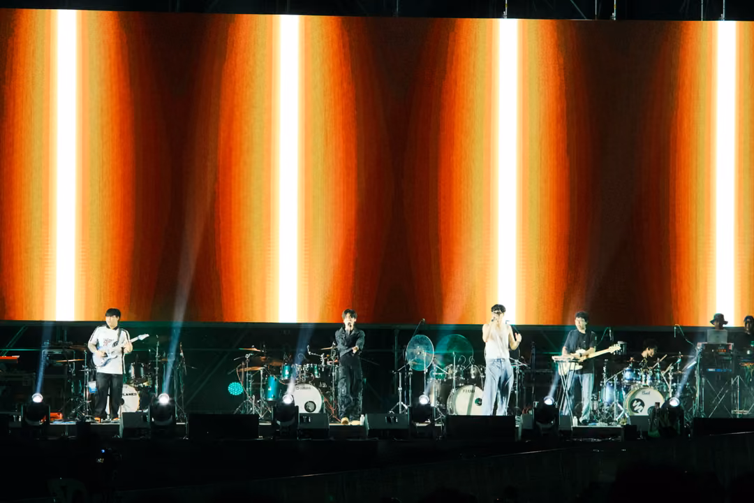 Stage with a screen of glowing orange vertical stripes, the band lined up in front, beams of light in the haze.