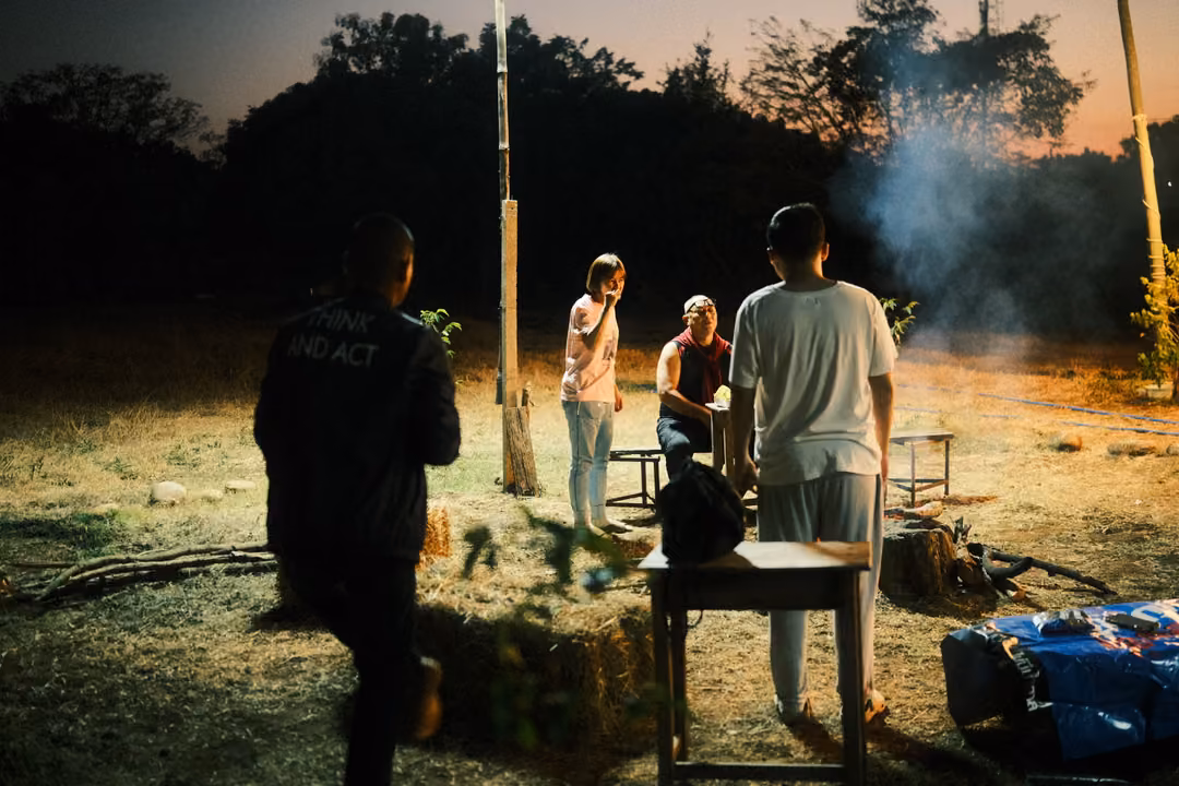 Group of people standing around a table and campfire on a dry lawn at dusk, smoke rising, trees behind