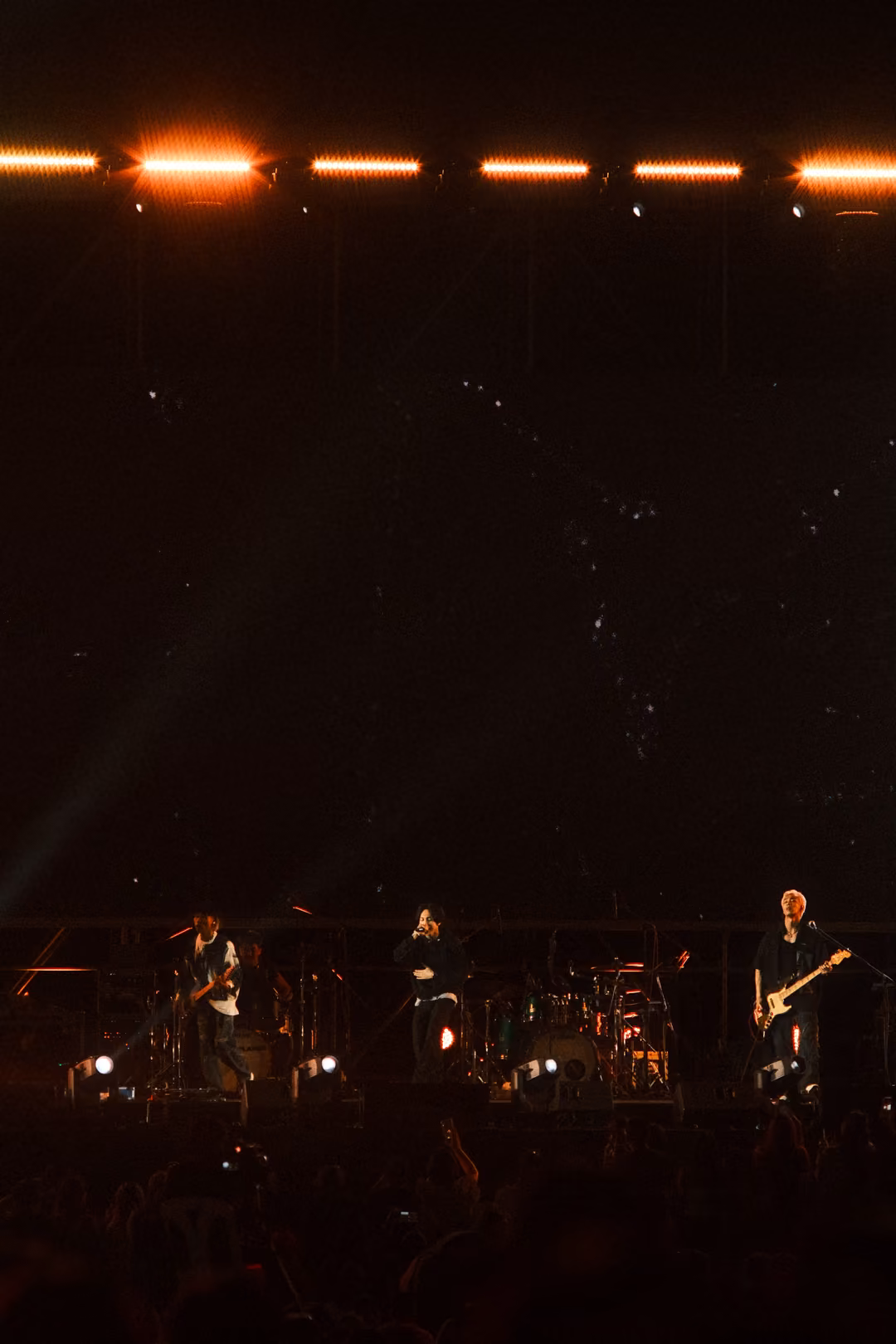 Vertical view of a dark stage with orange lights along the truss, three musicians performing in dim light.