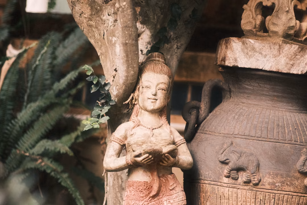 A carved figure holding a bowl, standing against a tree trunk beside a clay jar decorated with elephants.