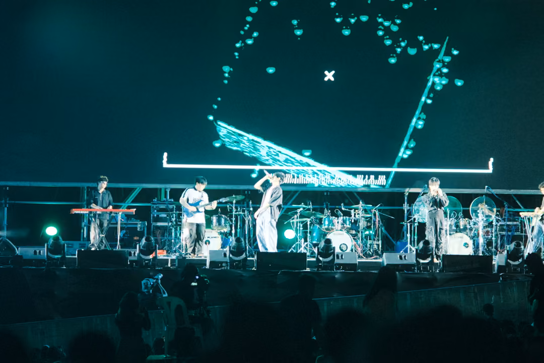 Stage with a screen showing turquoise shapes collapsing into a tilted plane, band performing below in green light.