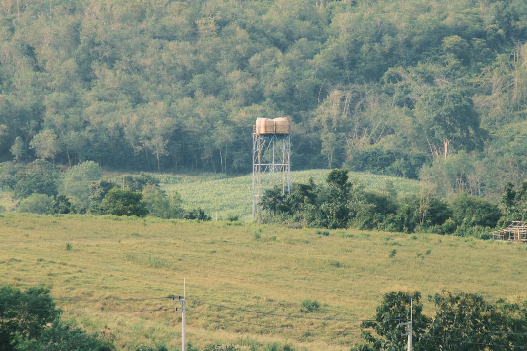A metal water tower standing among green fields and trees, with a forested hillside rising behind.