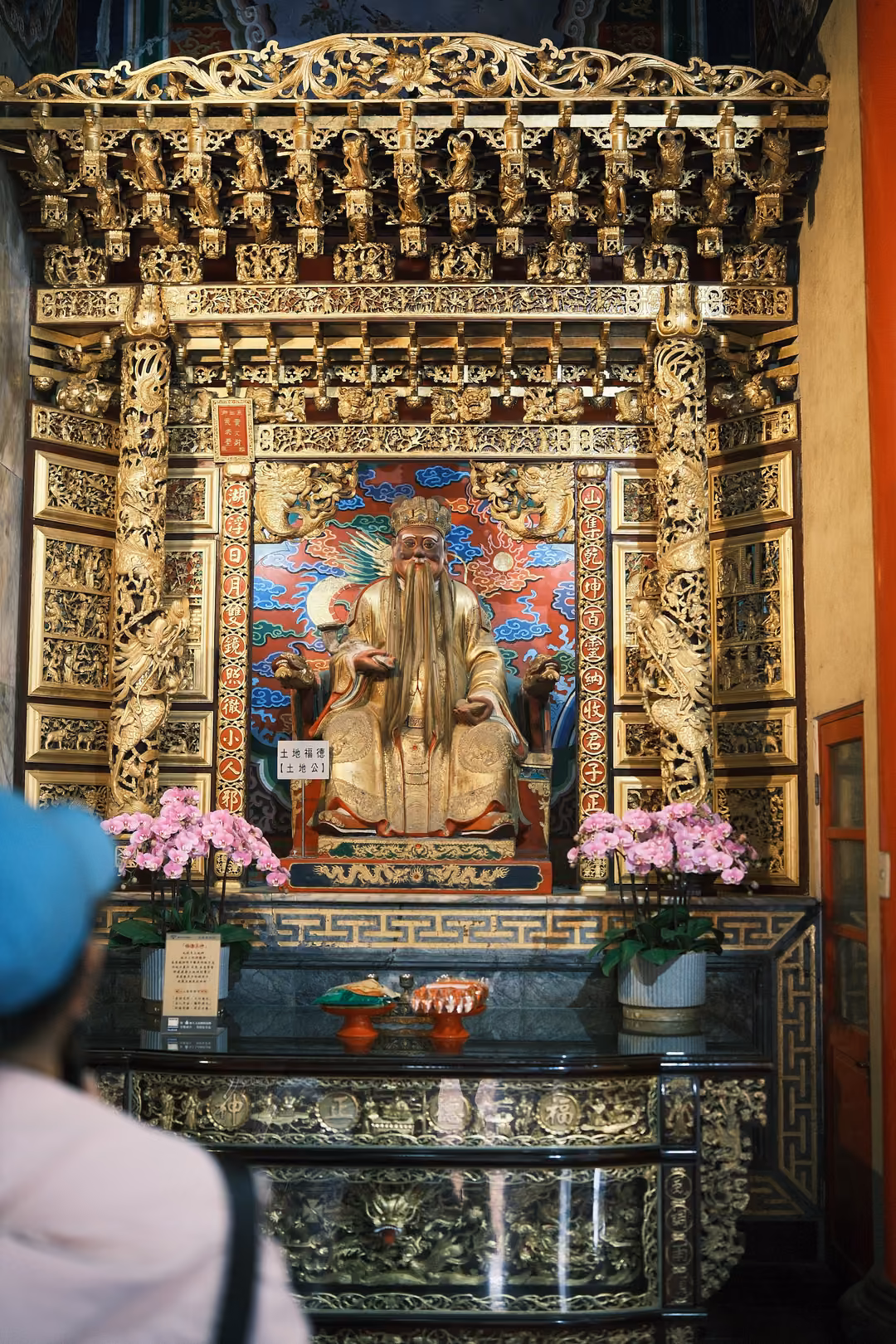 Gilded altar with a seated golden figure, pink orchids on either side, a visitor in a blue cap in the foreground