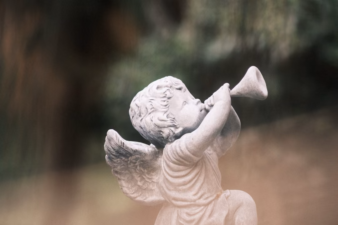 A small white cherub statue with wings blowing a trumpet, against a blurred garden background.