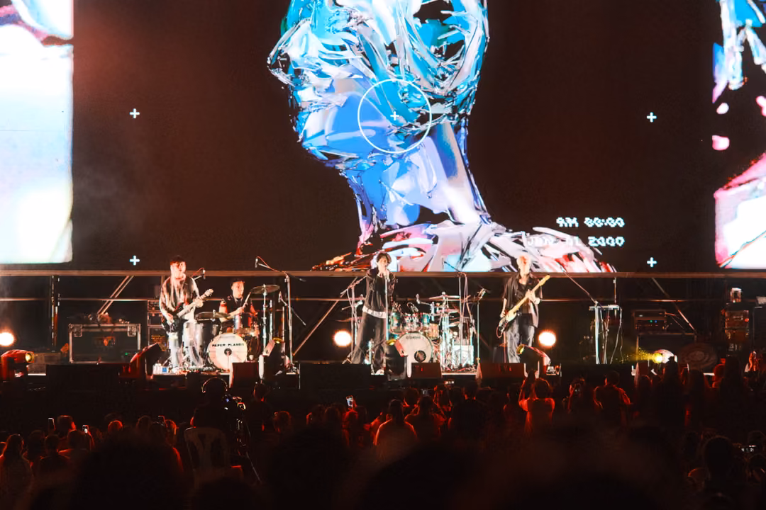 Band on an outdoor stage under a giant screen showing a chrome, glassy head in profile, crowd silhouetted below