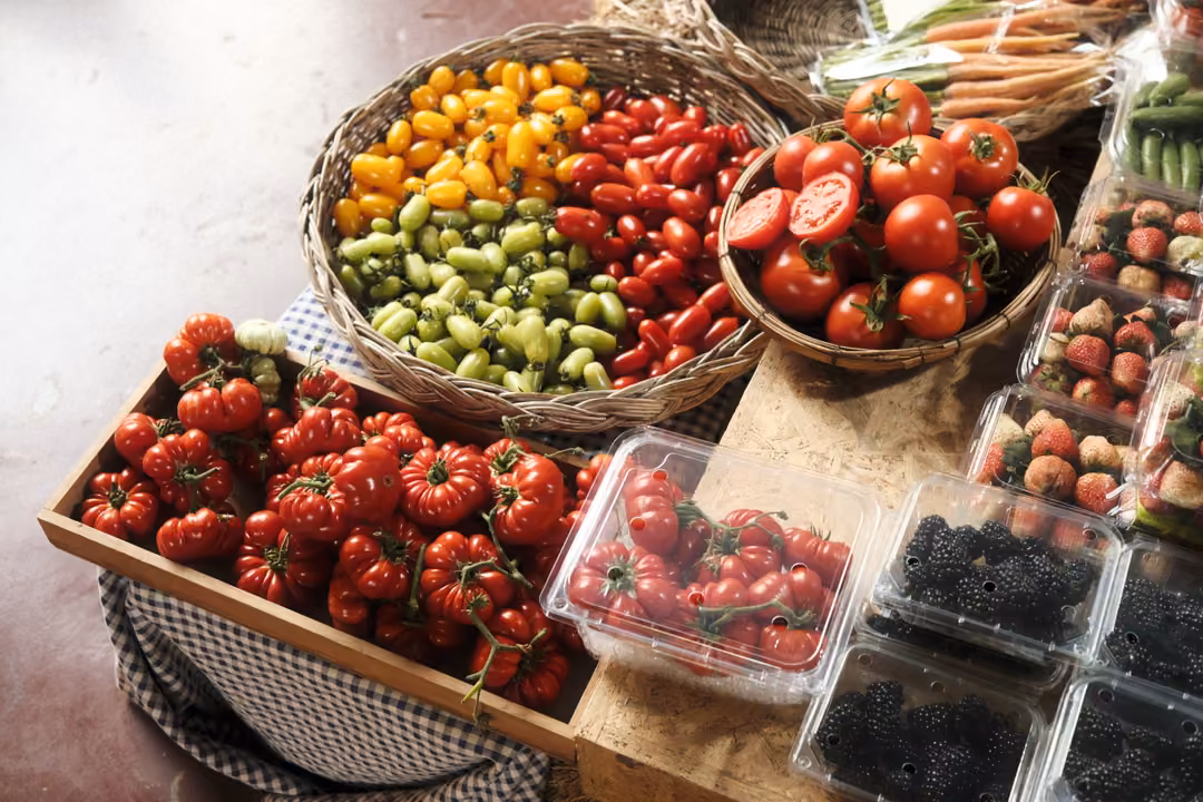 A market table with baskets of red and yellow tomatoes, punnets of berries and a checked cloth